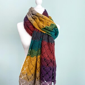Puede incluir: Una bufanda de ganchillo multicolor con un patr&oacute;n de encaje. La bufanda se muestra en un maniqu&iacute; blanco y presenta tonos de marr&oacute;n, amarillo mostaza, verde, morado y gris.