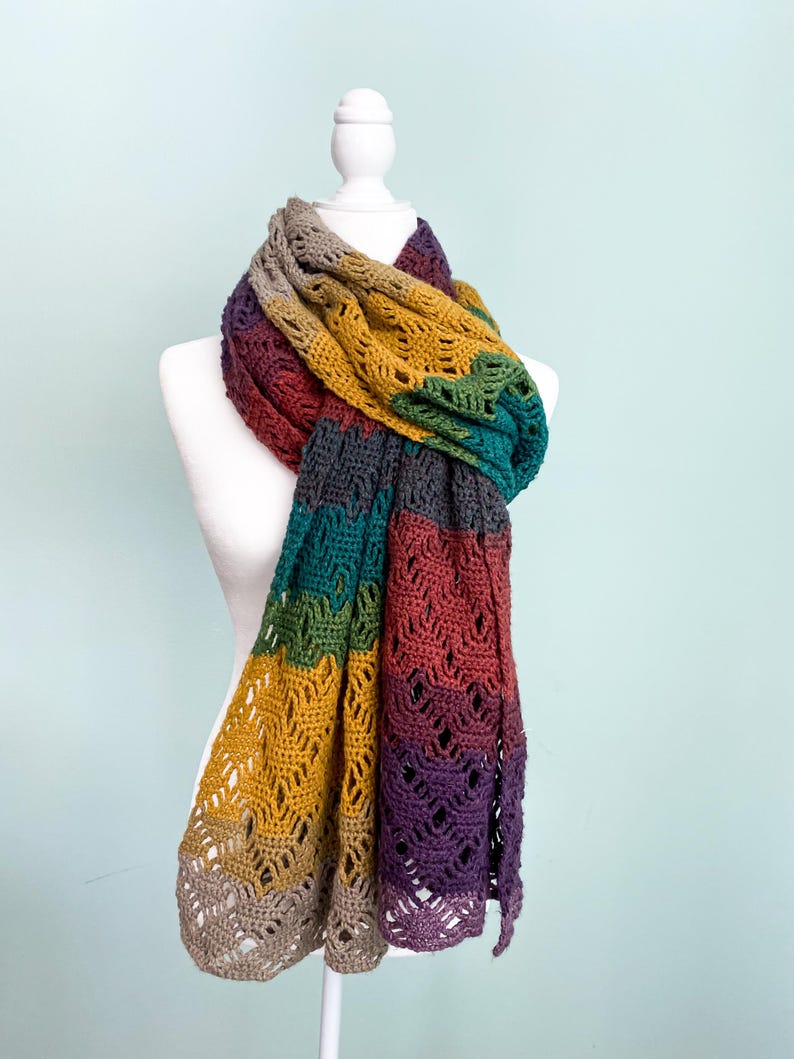 Puede incluir: Una bufanda de ganchillo multicolor con un patr&oacute;n de encaje. La bufanda se muestra en un maniqu&iacute; blanco y presenta tonos de marr&oacute;n, amarillo mostaza, verde, morado y gris.