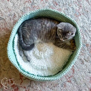 May include: A gray tabby cat resting in a light green crocheted cat bed with a white fluffy lining.