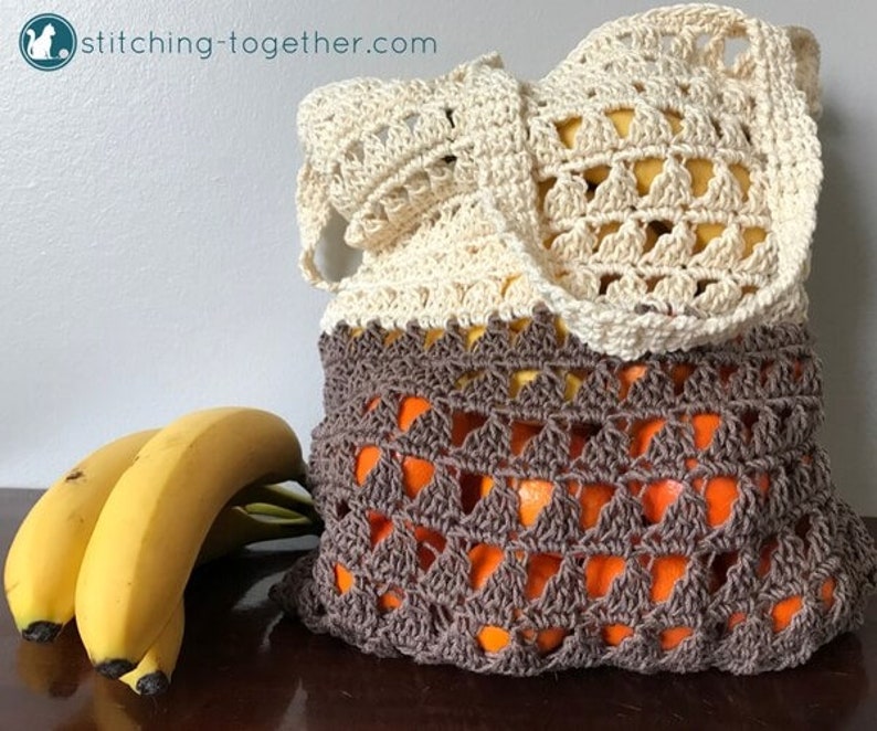 May include: A crocheted produce bag with a cream top and brown bottom. The bag is filled with oranges and is sitting on a wooden surface next to a bunch of bananas.