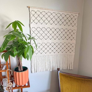 May include: Cream-coloured macrame wall hanging with a geometric diamond pattern and fringe. The wall decor is mounted on a wooden dowel. A potted plant with green leaves and a terracotta pot is in the foreground.