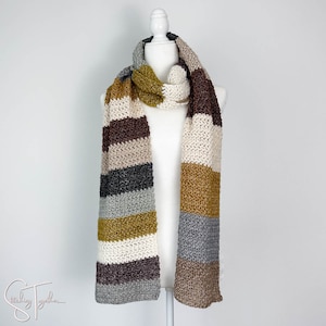 May include: A striped crocheted scarf in shades of brown, beige, gray, and gold. The scarf is displayed on a white mannequin.