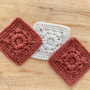 May include: Three crocheted squares, two in a rust color and one in white, with a floral design in the center of each square. The squares are arranged in a diagonal pattern on a wooden surface.