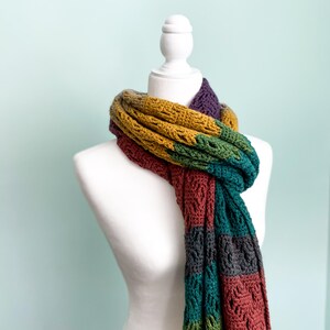Puede incluir: Una bufanda de crochet colorida con rayas de color amarillo mostaza, verde bosque, gris oscuro y burdeos. La bufanda est&aacute; drapeada alrededor de un maniqu&iacute; blanco.