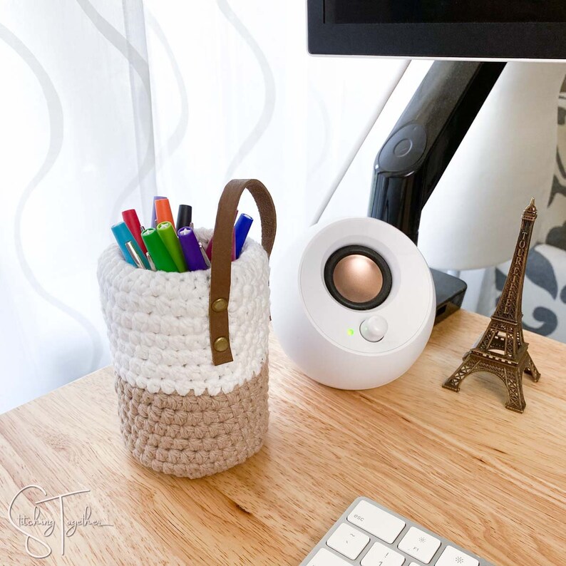 May include: A handmade, two-tone crochet pen holder with a brown leather handle. The top half is white, and the bottom is tan. The holder is filled with colorful pens. A white speaker and a miniature Eiffel Tower are also on the wooden desk.