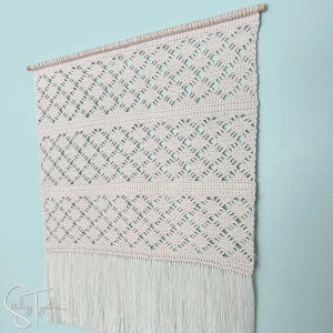 May include: Cream-colored macrame wall hanging with a geometric diamond pattern. The wall decor is suspended from a wooden dowel and features a fringe at the bottom. The piece is displayed against a light blue wall.