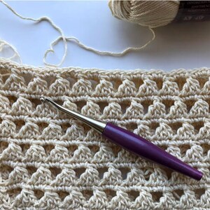 May include: A close-up of a crocheted fabric with a purple crochet hook. The fabric is a light beige color and has a repeating pattern of small triangles.