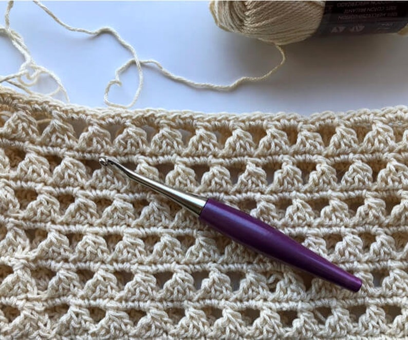 May include: A close-up of a crocheted fabric with a purple crochet hook. The fabric is a light beige color and has a repeating pattern of small triangles.