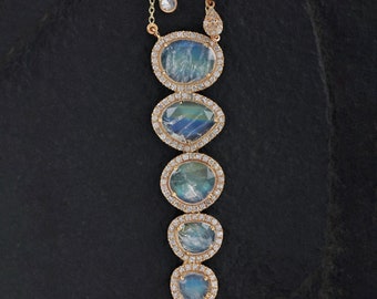 Può includere: Una collana in oro con cinque pietre ovali iridescenti allineate verticalmente, ciascuna circondata da un alone di piccoli diamanti bianchi. La catena include due ulteriori maglie rotonde con diamanti vicino alla chiusura.