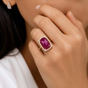 May include: A gold ring featuring a large, rectangular, faceted deep pink gemstone set in the center, surrounded by a halo of small, round, clear white stones. The band is thin and metallic gold, complementing the vibrant pink center stone.