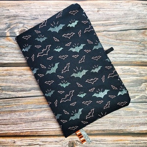 May include: A rectangular fabric book cover in black featuring a repeating pattern of silver glitter bats and thin white outlined bats. The cover includes a small black elastic loop on the side and two fabric ribbon bookmarks in orange and gray.