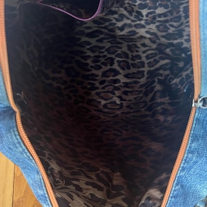 Puede incluir: Vista abierta de un bolso tote de mezclilla con ribete marr&oacute;n y forro con estampado de leopardo. El interior presenta un bolsillo con cremallera morada. El exterior de mezclilla azul contrasta con la tela de estampado animal.