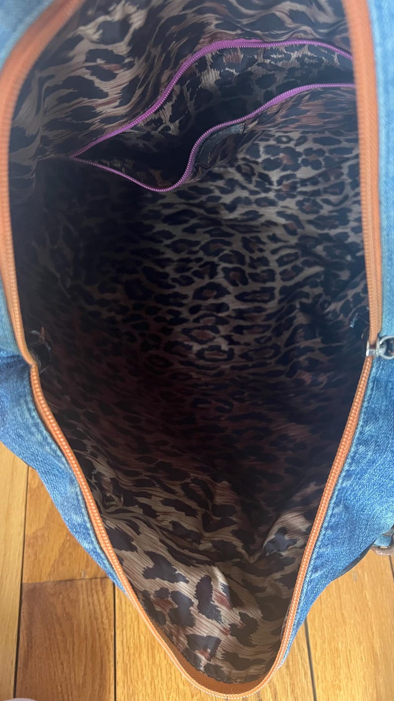 Puede incluir: Vista abierta de un bolso tote de mezclilla con ribete marr&oacute;n y forro con estampado de leopardo. El interior presenta un bolsillo con cremallera morada. El exterior de mezclilla azul contrasta con la tela de estampado animal.