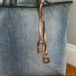 Puede incluir: Un bolso tote grande de mezclilla con un lavado azul claro. El bolso tiene un ribete y asas de color marr&oacute;n oscuro. Un dije de color cobre con las letras "DG" cuelga del asa. El bolso est&aacute; apoyado contra una pared blanca y un suelo de madera.