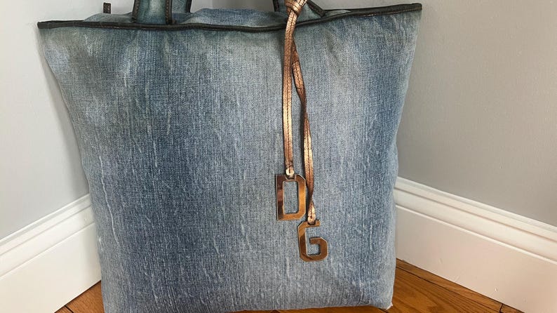 Puede incluir: Un bolso tote grande de mezclilla con un lavado azul claro. El bolso tiene un ribete y asas de color marr&oacute;n oscuro. Un dije de color cobre con las letras "DG" cuelga del asa. El bolso est&aacute; apoyado contra una pared blanca y un suelo de madera.