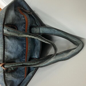 Puede incluir: Bolso tote de mezclilla azul con ribete y asas de cuero marr&oacute;n. El bolso cuenta con un cierre de cremallera y una franja central marr&oacute;n. Las asas est&aacute;n unidas con detalles de cuero marr&oacute;n.