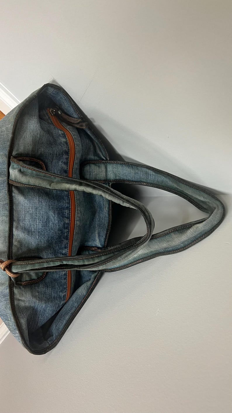 Puede incluir: Bolso tote de mezclilla azul con ribete y asas de cuero marr&oacute;n. El bolso cuenta con un cierre de cremallera y una franja central marr&oacute;n. Las asas est&aacute;n unidas con detalles de cuero marr&oacute;n.