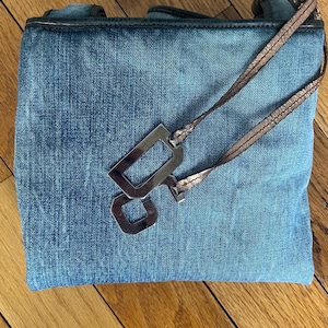 Puede incluir: Un bolso de mano de mezclilla azul con un dije rectangular plateado met&aacute;lico y una correa color cobre. El bolso tiene forma rectangular y una textura de mezclilla desgastada. La correa es delgada y parece ser de material met&aacute;lico.