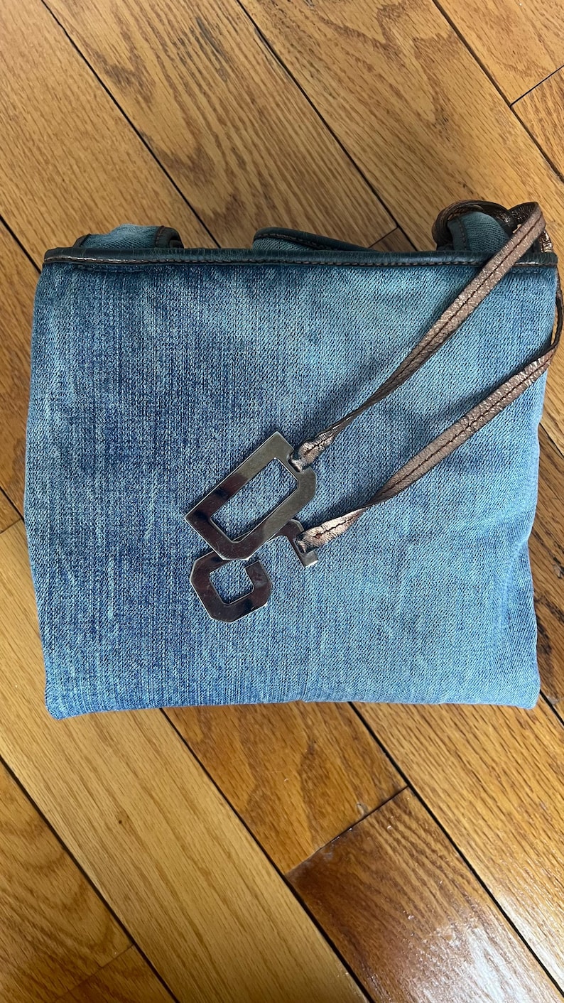 Puede incluir: Un bolso de mano de mezclilla azul con un dije rectangular plateado met&aacute;lico y una correa color cobre. El bolso tiene forma rectangular y una textura de mezclilla desgastada. La correa es delgada y parece ser de material met&aacute;lico.