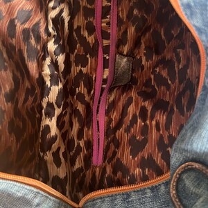 Puede incluir: Un bolso de mezclilla con forro de estampado de leopardo marr&oacute;n y negro. El bolso tiene una cremallera morada y un ribete naranja. El forro interior es visible, mostrando el estampado de leopardo.