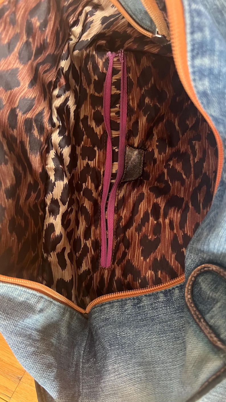 Puede incluir: Un bolso de mezclilla con forro de estampado de leopardo marr&oacute;n y negro. El bolso tiene una cremallera morada y un ribete naranja. El forro interior es visible, mostrando el estampado de leopardo.