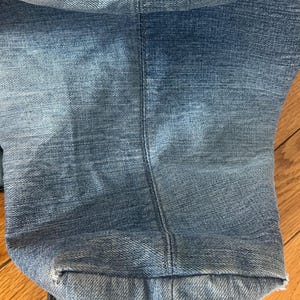 Puede incluir: Primer plano de unos vaqueros de mezclilla azul claro. La imagen muestra el dobladillo doblado y la costura de los vaqueros. La mezclilla tiene un aspecto descolorido, con zonas m&aacute;s claras y oscuras. Los vaqueros est&aacute;n sobre una superficie de madera.