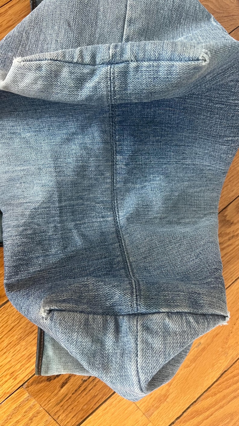 Puede incluir: Primer plano de unos vaqueros de mezclilla azul claro. La imagen muestra el dobladillo doblado y la costura de los vaqueros. La mezclilla tiene un aspecto descolorido, con zonas m&aacute;s claras y oscuras. Los vaqueros est&aacute;n sobre una superficie de madera.