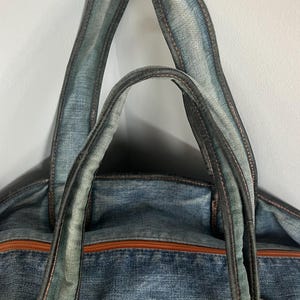 Puede incluir: Un bolso tote de mezclilla azul con ribetes y asas de cuero marr&oacute;n. El bolso presenta una cremallera marr&oacute;n y un detalle de cuero en el lateral. Las asas est&aacute;n reforzadas con costuras oscuras.