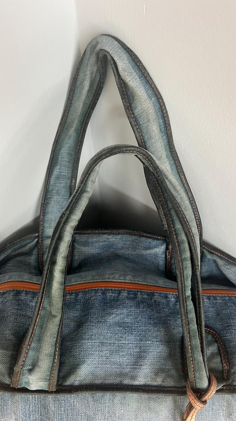 Puede incluir: Un bolso tote de mezclilla azul con ribetes y asas de cuero marr&oacute;n. El bolso presenta una cremallera marr&oacute;n y un detalle de cuero en el lateral. Las asas est&aacute;n reforzadas con costuras oscuras.