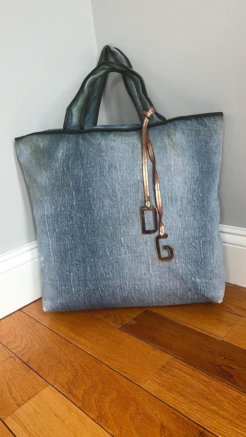 Puede incluir: Un bolso tote grande de mezclilla con un lavado azul descolorido. El bolso tiene asas verde oscuro y un ribete negro. Un dije dorado con las letras "DG" cuelga del asa. El bolso est&aacute; apoyado contra una pared gris claro.
