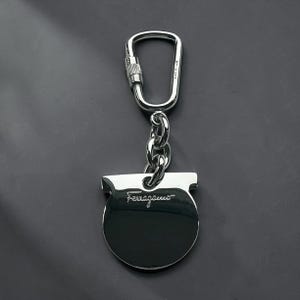 May include: A silver-colored keychain with a carabiner clip and a chain. The keychain has a circular tag with the brand name "Ferragamo" engraved on it. The background is a dark gray color.