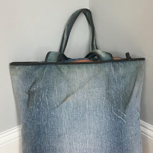 Puede incluir: Un bolso tote grande de mezclilla con dos asas. La bolsa est&aacute; hecha de tela de mezclilla azul con una apariencia descolorida. Las asas y el borde superior est&aacute;n recortados con un material m&aacute;s oscuro. La bolsa est&aacute; de pie contra una pared.