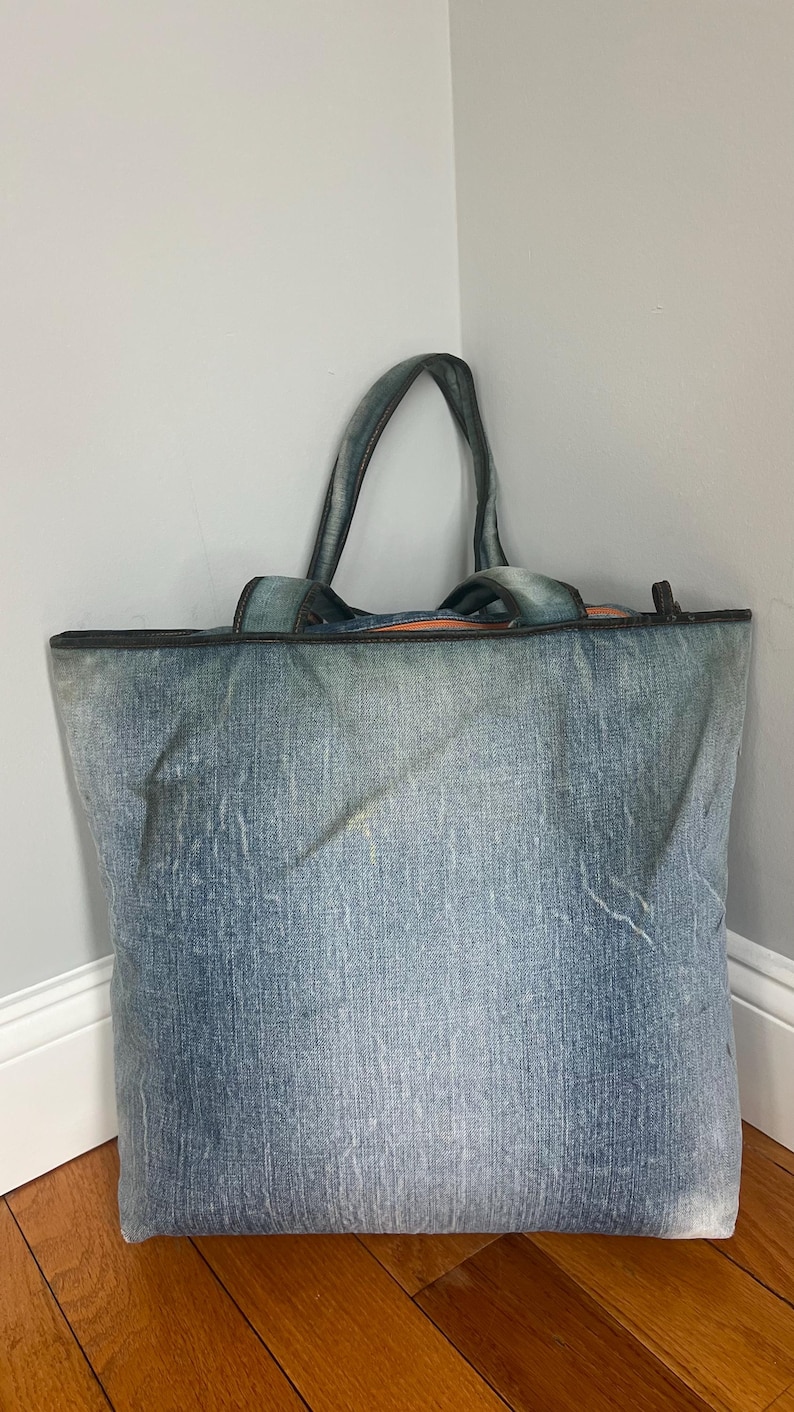 Puede incluir: Un bolso tote grande de mezclilla con dos asas. La bolsa est&aacute; hecha de tela de mezclilla azul con una apariencia descolorida. Las asas y el borde superior est&aacute;n recortados con un material m&aacute;s oscuro. La bolsa est&aacute; de pie contra una pared.