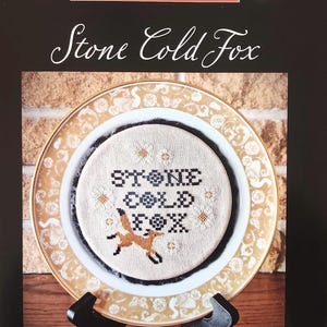 May include: A cross-stitch pattern titled "Stone Cold Fox" displayed on a decorative plate. The design features a fox and text in black and brown thread. The plate is on a black stand, with the text "The Artsy Housewife" at the top.