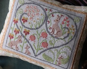 Op de afbeelding: Een kussen met een kruissteekpatroon met een hartvormig ontwerp met een konijn, bloemen en andere bosdieren. Het kussen ligt op een teal achtergrond met een chocoladekonijn en kwarteleieren.