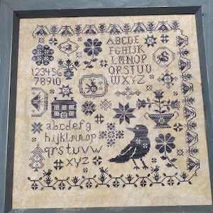 May include: A framed cross-stitch sampler with a black and white design. The sampler includes the alphabet, numbers, a small house, a bird, and a flower pot. The frame is a dark gray color.