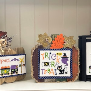 May include: Halloween-themed decorative wooden signs. The signs feature cross-stitch designs with pumpkins, trains, and houses. The signs are decorated with fall foliage, bows, and the phrases "Halloween Express", "Trick or Treat", and "Halloween Street".