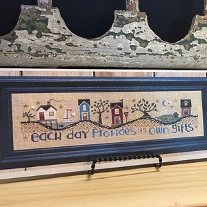 May include: A framed cross-stitch sampler with the text "each day provides its own gifts" and a depiction of houses on a hill. The sampler is displayed on a wooden shelf with a needlepoint pincushion on a wooden stand to the right.