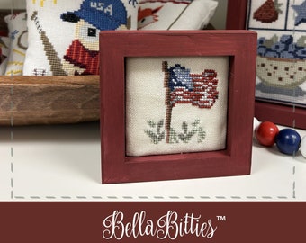 Può includere: Un ricamo a punto croce incorniciato di una bandiera americana con colori rosso, bianco e blu. La bandiera è su uno sfondo bianco con vegetazione verde alla base. La cornice è di colore rosso scuro. L'immagine è intitolata "Patriotic Flag" e fa parte della collezione "Bella Bitties".