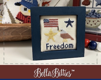 Può includere: Un ricamo a punto croce incorniciato che presenta una bandiera americana, una stella, una stella dorata e un'aquila con la parola "Freedom" sotto. La cornice è blu scuro e il punto croce è su tessuto bianco.