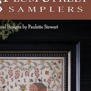 May include: A framed cross-stitch sampler titled "Tomato Sampler" by Plum Street Samplers. The design features a red brick building, a small house, and floral and berry motifs. The sampler is in a black frame.