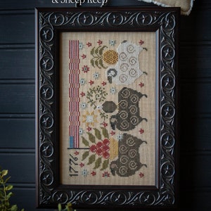 May include: Framed cross-stitch sampler titled "Flag Flock & Sheep Keep" by Plum Street Samplers. The design features sheep, floral elements, and a patriotic theme with the year 1776. The frame is dark brown with decorative carvings.