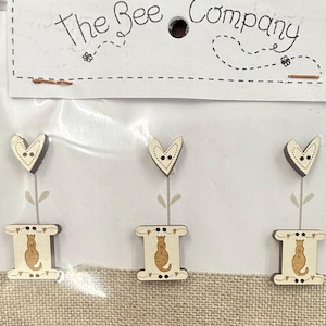 May include: Three wooden buttons with a floral design. Each button features a heart-shaped flower with a dotted face and a spool-shaped base with a cat silhouette. The top of the card reads "The Bee Company".