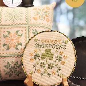 Puede incluir: Un bastidor de bordado de punto de cruz con un diseño de trébol y el texto "Collect Shamrocks". El bastidor se muestra sobre un soporte de madera, con un cojín decorativo en el fondo. El texto "Tiny Tarts" está en la parte superior.