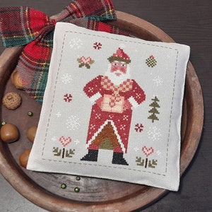 May include: A white cross-stitch sampler featuring a red and white Santa Claus figure in a snowy winter scene. The sampler is framed with a grey border and is displayed on a wooden surface.