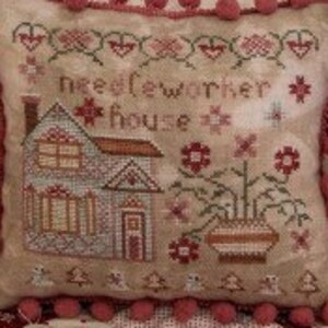 May include: A small, white pillow with a red and white embroidered design that says "Needleworker House". The pillow has a red pom-pom trim. A white snowman with a green and white striped scarf and red earmuffs is standing next to the pillow.