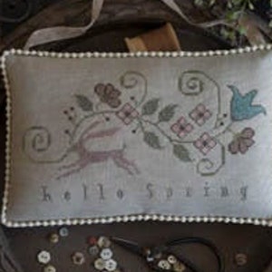 May include: A white pillow with a cross-stitch design featuring a floral motif and the text "Hello Spring". The pillow is on a wooden tray with buttons and a pair of scissors.