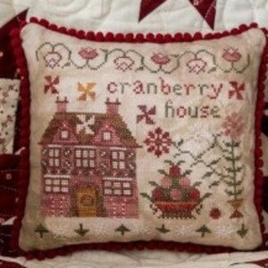 May include: A white fabric snowman with a green and white striped scarf and a red pom-pom hat. The snowman is holding a green fabric leaf in its hand. The snowman is sitting on a white fabric background with a red and white quilt behind it. The quilt has a star pattern. A small pillow with a red border and a cross-stitch design of a house with the text "cranberry house" is in the foreground.