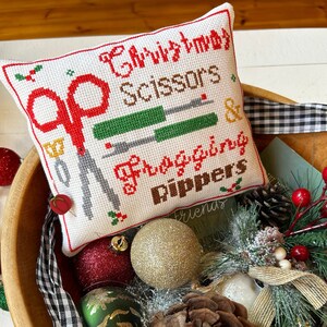 Può includere: Un cuscino bianco ricamato a punto croce con le parole "Christmas Scissors & Frogging Rippers" in rosso e marrone. Il cuscino presenta immagini di forbici e taglia-cuci. Il cuscino è in una ciotola di legno con decorazioni natalizie e pigne.
