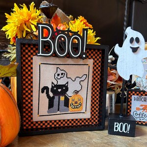May include: A black framed cross-stitch with a black cat, a ghost, and a jack-o-lantern. The frame has a black and orange checkered border. The frame is sitting on a wooden surface with a wooden ghost and a sign that says "BOO!" in front of it.